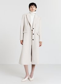 Cappotto lungo color beige chiaro realizzato in un tessuto morbido, con colletto a revers, grandi bottoni neri e tasche laterali. Indossato sopra un dolcevita bianco.