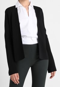 Black, open-front cardigan in soft fabric, long sleeves. Paired with a white collared shirt and dark slim-fit pants. Minimalist design.