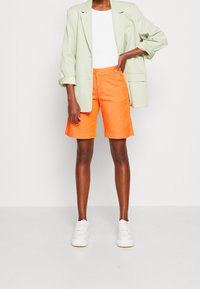 Světle zelený oversized blazer, bílý tričko a jasně oranžové šortky. Modelka má na sobě bílé tenisky. Módní vrstvený outfit s živými barvami.