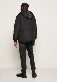 Giubbotto piumino nero con cappuccio, caratterizzato da un logo sul retro, maniche arricciate e una vestibilità oversized, abbinato a pantaloni in denim scuro e stivali neri.