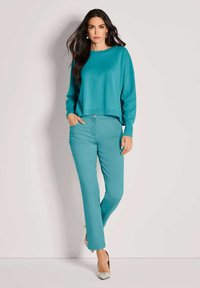 Türkiser Pullover mit rundem Halsausschnitt und langen Ärmeln, kombiniert mit türkisen schmal geschnittenen Hosen und gemusterten High Heels.