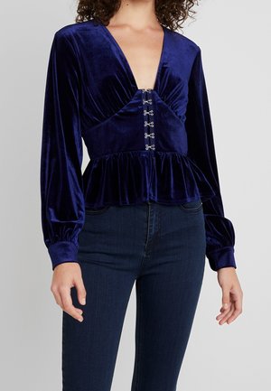 Kobieta ubrana w ciemnoniebieską aksamitną bluzkę z długim rękawem, z zapięciem z haczyków i oczek z przodu oraz z baskinką, w połączeniu z ciemnoniebieskimi dżinsami.