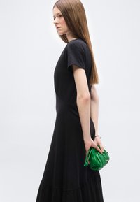 Γυναίκα με ίσια μακριά μαλλιά φοράει κοντομάνικο μαύρο φόρεμα και κρατά μια μικρή πράσινη τσάντα πίσω από την πλάτη της, κοιτάζοντας προς τα αριστερά.