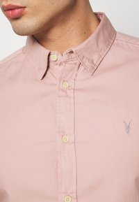 Camisa de manga larga de color rosa claro con cuello estructurado, botones amarillos y un pequeño emblema en relieve. Tejido texturizado, diseño casual.