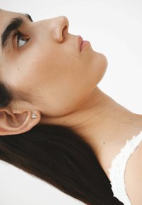 Perfil de una persona con cabello largo y oscuro, que lleva un pequeño arete brillante. El tono de piel es medio. El fondo es blanco liso.