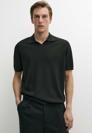 Junger Mann mit hellbraunen Haaren, der ein schwarzes, kurzärmliges, geripptes Poloshirt und schwarze Hose trägt, steht vor einem schlichten weißen Hintergrund.