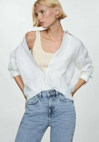 White linen shirt, open at the front, over a light beige tank top. Light blue high-waisted jeans with a distressed look. Simple design.
