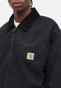 Άτομο που φοράει μαύρο μπουφάν Carhartt με φερμουάρ και τσέπη στο στήθος με κίτρινο σήμα στο αριστερό πλευρό.
