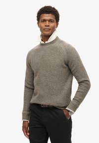 Pull en maille texturée marron clair avec un col montant, des poignets côtelés, et une chemise blanche en dessous. Assorti avec un pantalon sombre et une montre.