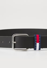 Schwarzer Ledergürtel mit einer silbernen rechteckigen Schnalle; verfügt über einen blauen Patch mit roten und weißen Streifen, beschriftet mit "TOMMY JEANS."