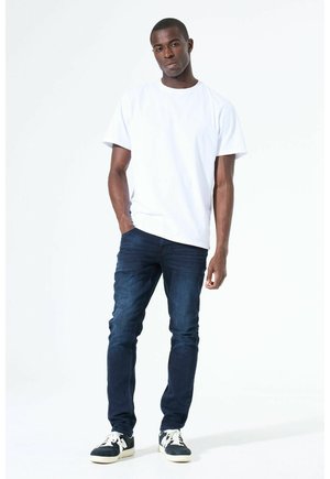 Mann steht vor weißem Hintergrund, trägt ein weißes T-Shirt, dunkelblaue Jeans und schwarz-weiße Turnschuhe, mit einer Hand in der Tasche.