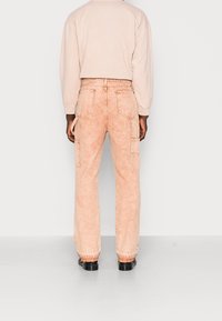 Persona vestida con pantalones cargo naranja claro y una camiseta de manga larga beige, de pie con la espalda hacia la cámara en un fondo liso.