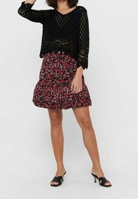 Schwarzes gehäkeltes Oberteil mit V-Ausschnitt und lockeren Ärmeln, kombiniert mit einem ausgestellten, floral gemusterten Rock in Schwarz, Pink und Lila. Schwarze Absatzsandalen.