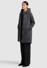 Donna in piedi che indossa un lungo parka con cappuccio grigio scuro e tasche frontali, pantaloni neri a gamba larga e stivali neri con tacco su sfondo chiaro.