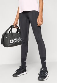 Zwarte leggings van rekbare stof, gecombineerd met een lichtroze top en zwarte sneakers. Er wordt een Adidas weekendtas met witte logo-accenten vastgehouden.