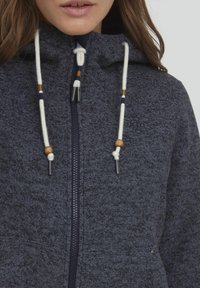 Haut à capuche bleu marine en tissu texturé, avec des cordons ajustables en bois, et une fermeture éclair à l'avant. Dispose de poches latérales.