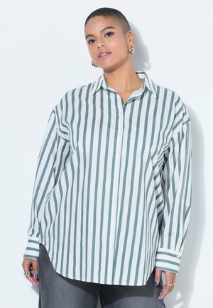 Osoba s krátkými vlasy nosí nadměrnou bílou košili s vertikálními šedými pruhy, tmavé kalhoty, zlaté kruhové náušnice a modrý lak na nehty.