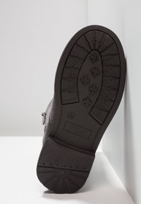 Semelle d'une botte noire avec des profondes rainures, logo "Xti", marquage taille 29, positionnée verticalement sur un fond blanc et gris.