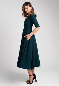 Turkusowa sukienka do kolan z krótkimi rękawami, dopasowanym górnym partem, rozkloszowanym dołem i bocznymi kieszeniami. Połączona z czarnymi sandałami na wysokim obcasie.