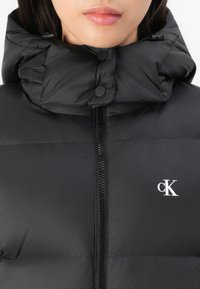 Musta puhviviesti korkealla kauluksella, painonapeilla ja vetoketjulla. Edessä valkoinen "cK" logo. Tasainen ja kiiltävä pinta.