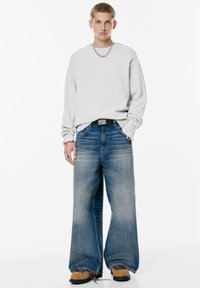 Hanorac oversized gri deschis cu mâneci lungi, purtat cu blugi albaștri cu picioare largi. Accesoriile includ o brățară cu lanț și o curea neagră.