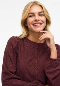 Vinrød tekstureret bluse med lange puffede ærmer og rund halsudskæring, dekoreret med en række knapper foran og indviklede mønsterdetaljer.