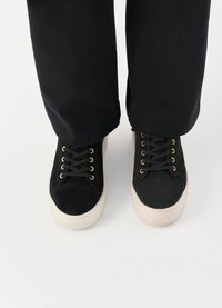 Schwarze Wildleder-Sneaker mit abgerundeter Spitze, weißen Gummisohlen, schwarzen Schnürsenkeln und goldenen Ösen, kombiniert mit lockeren schwarzen Hosen.
