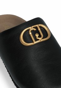 Zwarte leren slide sandaal met een groot hardware logo in goudkleur op de bovenkant en een gladde voering aan de binnenzijde. Eenvoudig, rond ontwerp.