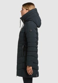 Cappotto piumino nero con colletto alto, cappuccio regolabile e polsini a coste. Caratterizzato da una tasca laterale con zip e toppa con logo sulla manica.