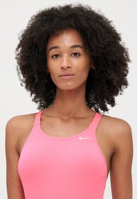 Costume da bagno sportivo rosa con spalline sottili, realizzato in tessuto morbido. Presenta un piccolo logo bianco Nike nell'area sinistra del petto.