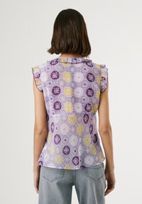 Lila mönstrad ärmlös topp med volanger vid axlarna, som har gula och vita blommönster på ett strukturerat tyg.