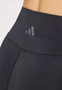 Aukštos juosmeninės juodos sportinės legginsai, pagaminti iš lygaus, elastingo audinio, su tekstūruota juosmeniu ir atspindinčiu logotipu priekyje.