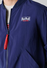 Marineblauw jack met ritssluiting, voorzien van het Red Bull Racing-logo op de borst en een rode ritstrekkker, gedragen over een wit shirt met rode en blauwe strepen.