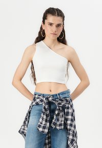 Witte geribbelde crop top met één schouderband, gecombineerd met hoge blauwe jeans en een geknoopte geruite blouse rond de heupen.