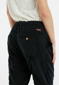 Zwarte cargobroek met een elastische tailleband, zijzakken en twee achterzakken. Bevat een bruine knoop en een klein stoffen label.