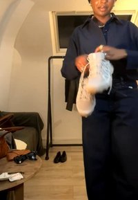Personne en vêtements foncés tenant des bottes blanches dans une pièce avec un porte-manteau, des chaussures au sol et une petite table avec des lunettes de soleil.