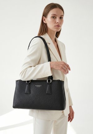 Femme en tailleur blanc tenant un sac à main en cuir noir Diana&Co Firenze sur l'épaule, sur fond uni.