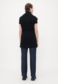 Person med kort hår iført en svart ermeløs strikket vest med belte, mørkeblå jeans og svarte klumpete sko, stående vendt bort.