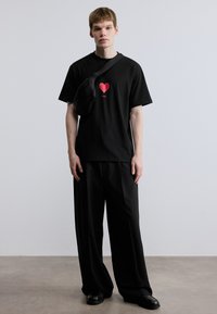 Μαύρο κοντομάνικο μπλουζάκι με κόκκινο γραφικό καρδιάς στο κέντρο, συνδυασμένο με φαρδιά μαύρη παντελόνια και μια μικρή μαύρη τσάντα ώμου.