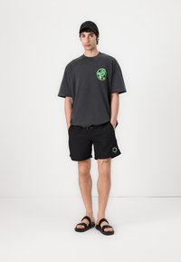 Løs pasform mørkegrå t-shirt med et grønt grafisk motiv, parret med sorte shorts med et hvidt logo. Båret med sorte sandaler.