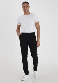 Schwarze, maßgeschneiderte Hose aus leichtem Stoff, schmaler Schnitt, mit Knopfverschluss. Kombiniert mit einem schlichten weißen Kurzarmhemd und weißen Turnschuhen.