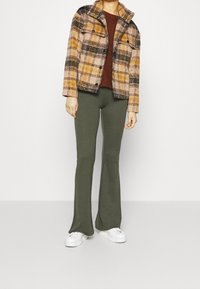 Veste à carreaux beige, orange et noir avec un col, associée à un pantalon évasé vert olive. Des baskets blanches complètent la tenue.