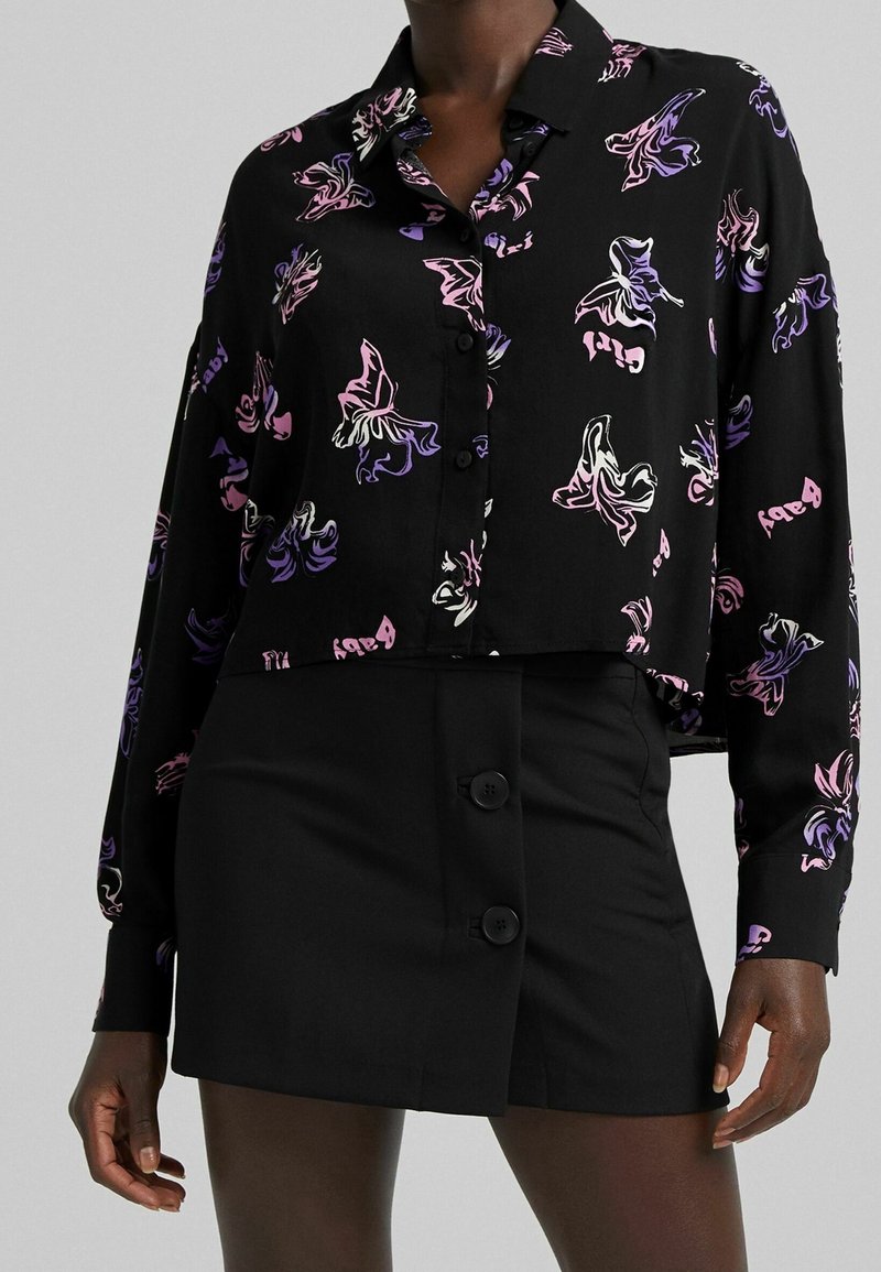 Femme portant une jupe noire boutonnée et une chemise noire à manches longues avec des imprimés de papillons roses et violets sur fond clair.