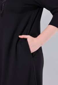Schwarzes Kleid mit Rundhalsausschnitt, drei Viertelärmeln, Seitentaschen und einer glatten, weichen Textur. Mit einem subtilen, glänzenden Detail akzentuiert.