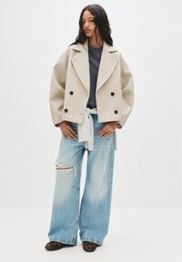 Cappotto in misto lana beige a doppio petto con grandi bottoni neri, abbinato a una maglia scura, jeans strappati chiari e scarpe con stampa animale.