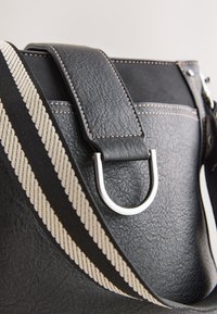 Gros plan d’un sac en cuir noir texturé avec anneau en D argenté et large bandoulière rayée noire et blanche.