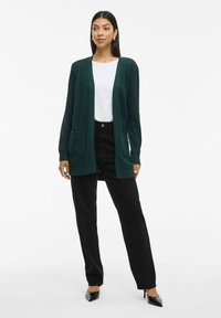 Temno zelena jopica z teksturirano površino, odprto spredaj, dolgi rokavi in sprednji žep, v kombinaciji s črnimi hlačami in koničastimi petami.