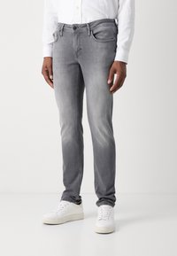 Vaqueros de denim gris con un corte ajustado, un ligero desvanecimiento y un diseño de cinco bolsillos, combinados con zapatillas blancas sobre un fondo sencillo.