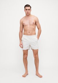 Svetlo béžové plavky s elastickým pásom, bočnými vreckami a hladkým povrchom. Nosené mužským modelom, ktorý stojí naboso.