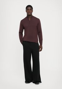 Pull rouge à manches longues avec un col zippé quart, associé à un pantalon large noir et des chaussures noires. Texture tricot lisse.